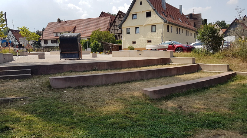 Dorfplatz mit treppenartigen Stufen am Flussufer, ein Strandkorb, Parkplatz und Häuser im Hindergrund.