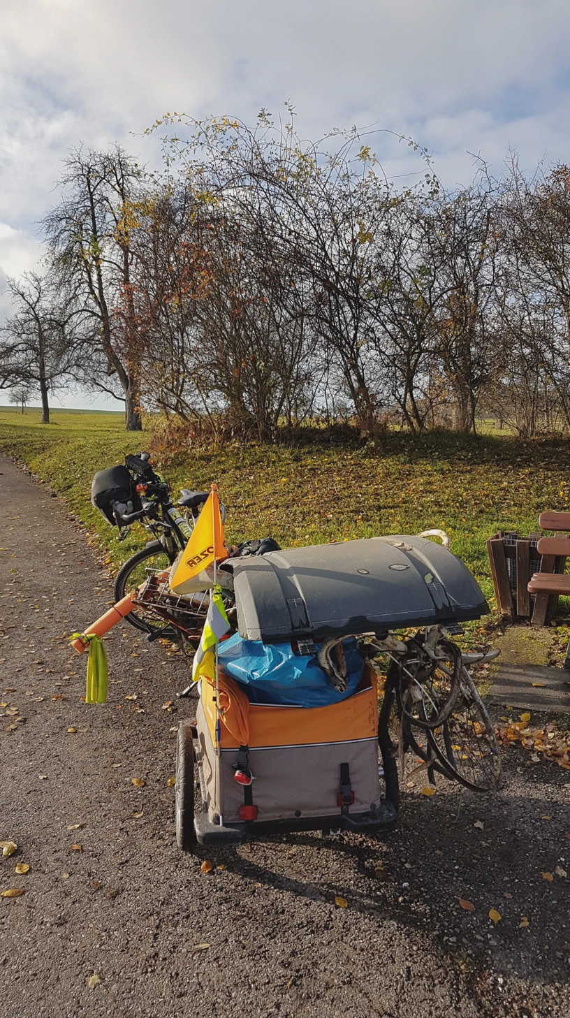 Gespann aus Fahrrad und beladenem Anhänger und oben auf dem Anhänger liegt noch die Radabdeckung eines Lkw ohne Sicherung.