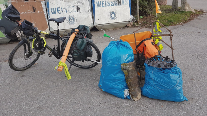 Gespann aus Fahrrad und Fahrradhänger. Vor dem Anhänger stehen zwei volle Müllsäcke, die Egge, eine Radkappe eines Autos und der Rest eines Steinrohrs.
