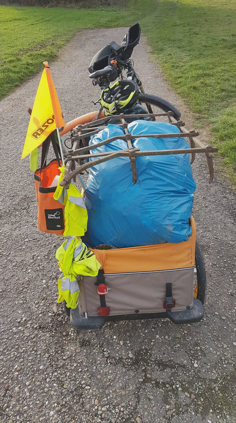 Gespann aus Fahrrad und Fahrradanhänger mit einem Müllsack beladen und oben drauf liegt die Egge.