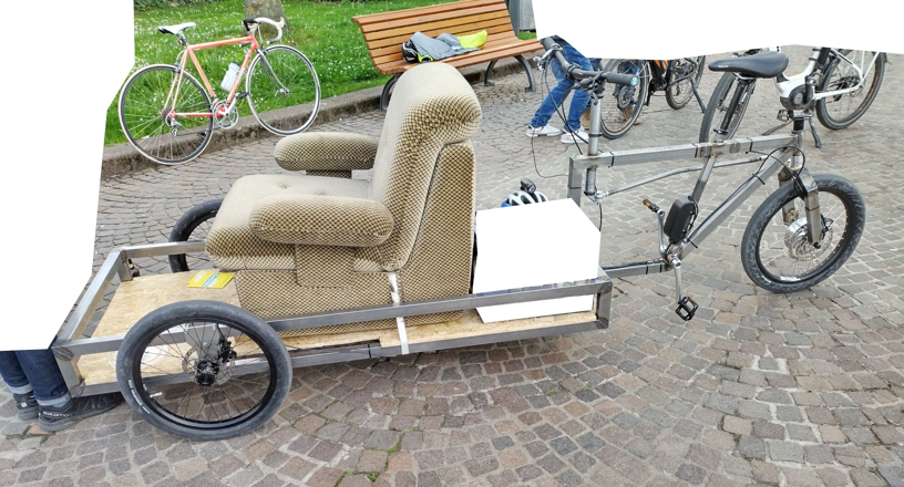 Lastenrad mit Sessel. Ein Lastenrad mit Ladefläche vorne, auf dem ein Sessel mit Stoffbezug befestigt ist.