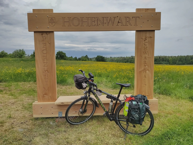 Ein Fahrrad steht vor dem Hohenwarter Fotorahmen und in der Lenkertasche sitzt der kleine Elefant. Außerhalb vom Rahmen schöne Landschaft und grau bewölkter Himmel.