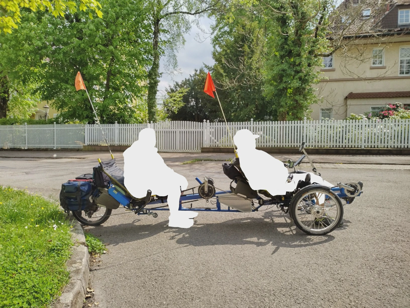 Liegedreirad-Tandem für zwei Menschen von der Seite fotografiert, wobei beide Menschen ausgeschnitten wurden, um sie unkenntlich zu machen.