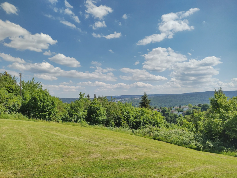 Landschaft mit Wiese im Vordergrund, Bäumen in der Mitte, Hügelkette mit Dörfern im Hintergrund und schönem blauen Himmel mit weißen Wolken.
