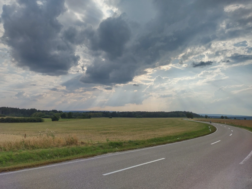 Schlängelnde Landstraße, Felder, Himmel mit sichtbaren Sonnenstrahlen.
