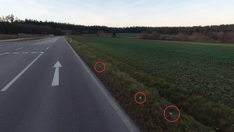 Landstraße mit Feldern rechts und links. Rechts im Bild sind im Gras drei Markierungen im Bild. Dort liegen jeweils Geldscheine.