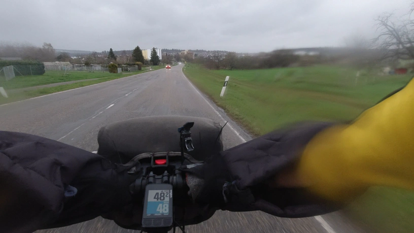 Ausschnitt aus einem nach vorne ausgerichteten Video beim Fahrrad fahren. Landstraße, die ich bergab befahre, links und rechts Felder. In der Bildmitte der Fahrradcomputer am Lenker, der oben die Geschwindigkeit mit 48,2 km/h anzeigt und darunter und blau hinterlegt den Puls mit 48.